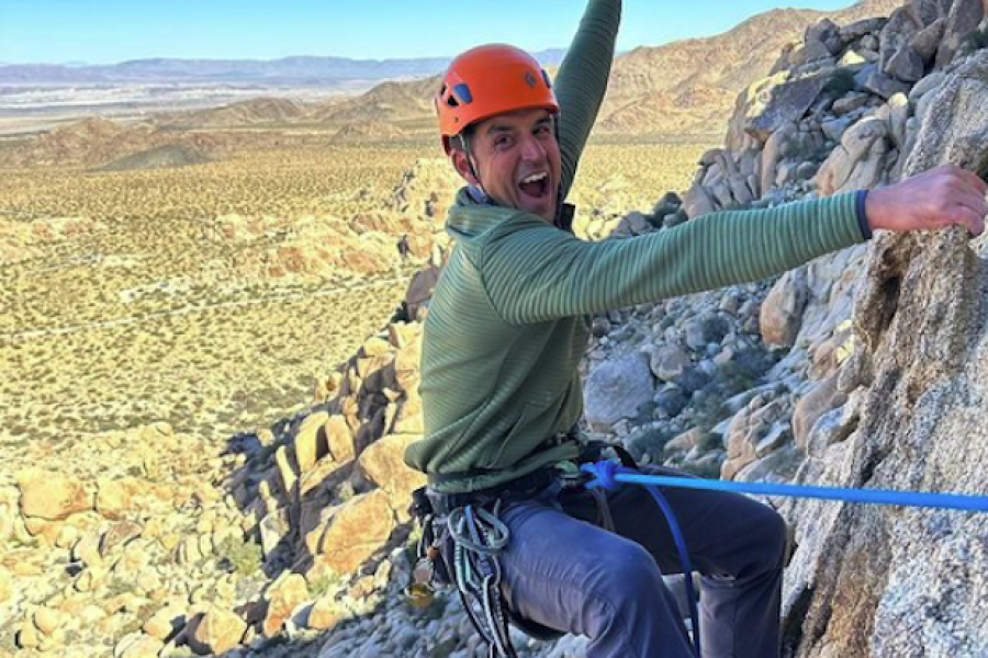 A man with orange climbing helmet, green ribbed long sleeve, and grey pants in rock climbing harness hangs on a rock with right hand with left hand raised in the air during Joshua Tree guide-led day trip in southern California.
